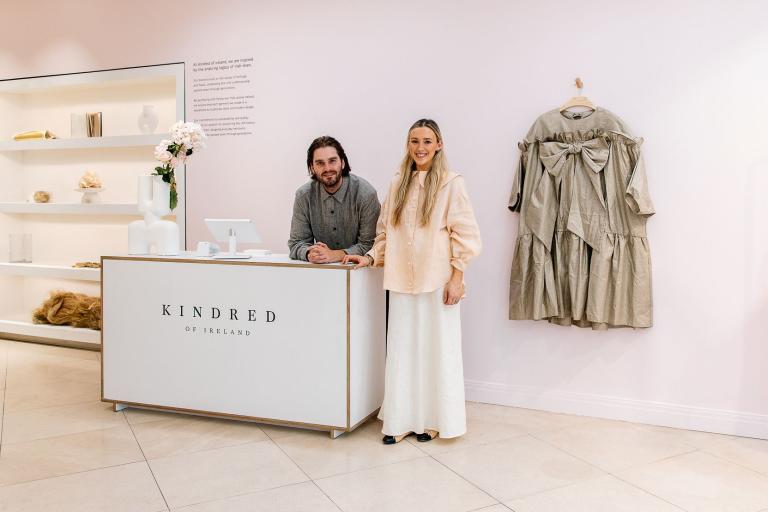 the owners of kindred of ireland standing in their new store, with clothes hanging up and accessories on shelves